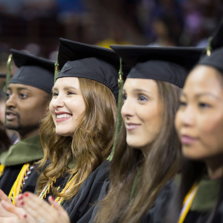 Students in their cap and gown sit in a row at graduation. They are smiling.