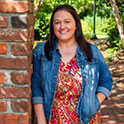 Brittany is pictured from the waist up wearing a colorful dress. She has brown hair, fair skin, and is smiling. 