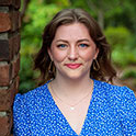 Sam is pictured from the shoulders up wearing a blue dress. She has fair skin and is smiling. 