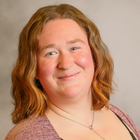 Professional headshot of a smiling person with shoulder-length wavy light brown hair, wearing a maroon knit cardigan over a black top and a delicate silver necklace. They are turned slightly to one side while looking directly at the camera. The background is a softly blurred neutral gray, giving the portrait a warm and approachable appearance.