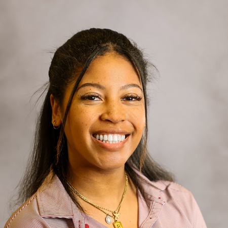 Professional headshot of a smiling person with long dark hair styled half up and half down wearing a light gray and garnet striped collared polo shirt with a university logo. The person is facing the camera and is wearing small hoop earrings and layered gold necklaces. The photo is set against a soft gray studio background with warm, even lighting, creating a friendly, approachable, and professional appearance.