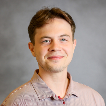 Professional headshot of a smiling person with short brown hair wearing a light gray and maroon striped collared polo shirt. The person is facing the camera against a soft gray studio background with even lighting, creating a polished and approachable appearance.