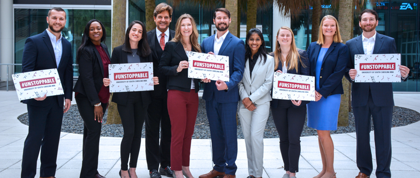 IMBA students standing in the Moore School courtyard, under palmetto trees and holding sings announcing that the IMBA program has been ranked no. 1 by U.S. News and World Report 