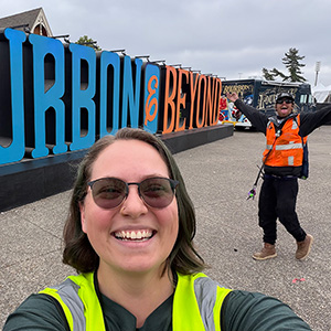 Woman in sunglasses and yellow utility vest poses before 'Bourbon and Beyond' festival sign with playful coworker gesturing in background. 